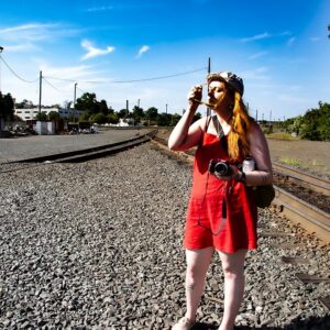 Jade Alexis Wollin standing between railroad tracks holding a camera in one hand and putting on sunglasses with the other.